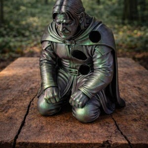 May include: A metallic, dark green and bronze-toned sculpture of a kneeling figure with long hair, wearing a hooded cloak and tunic. The figure has three circular, hollow voids in the chest and shoulder area, with a somber facial expression.