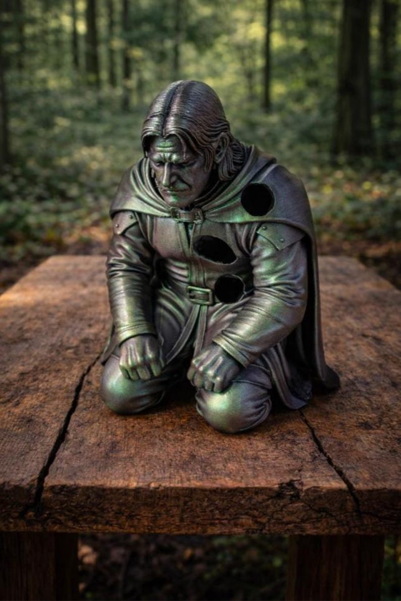May include: A metallic, dark green and bronze-toned sculpture of a kneeling figure with long hair, wearing a hooded cloak and tunic. The figure has three circular, hollow voids in the chest and shoulder area, with a somber facial expression.