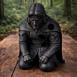 Op de afbeelding: Een zwart beeld van een knielende figuur met lang haar en een cape. Het beeld staat op een houten oppervlak, met een vage bosachtergrond. De figuur draagt een leren outfit met een riem en pantserachtige details.