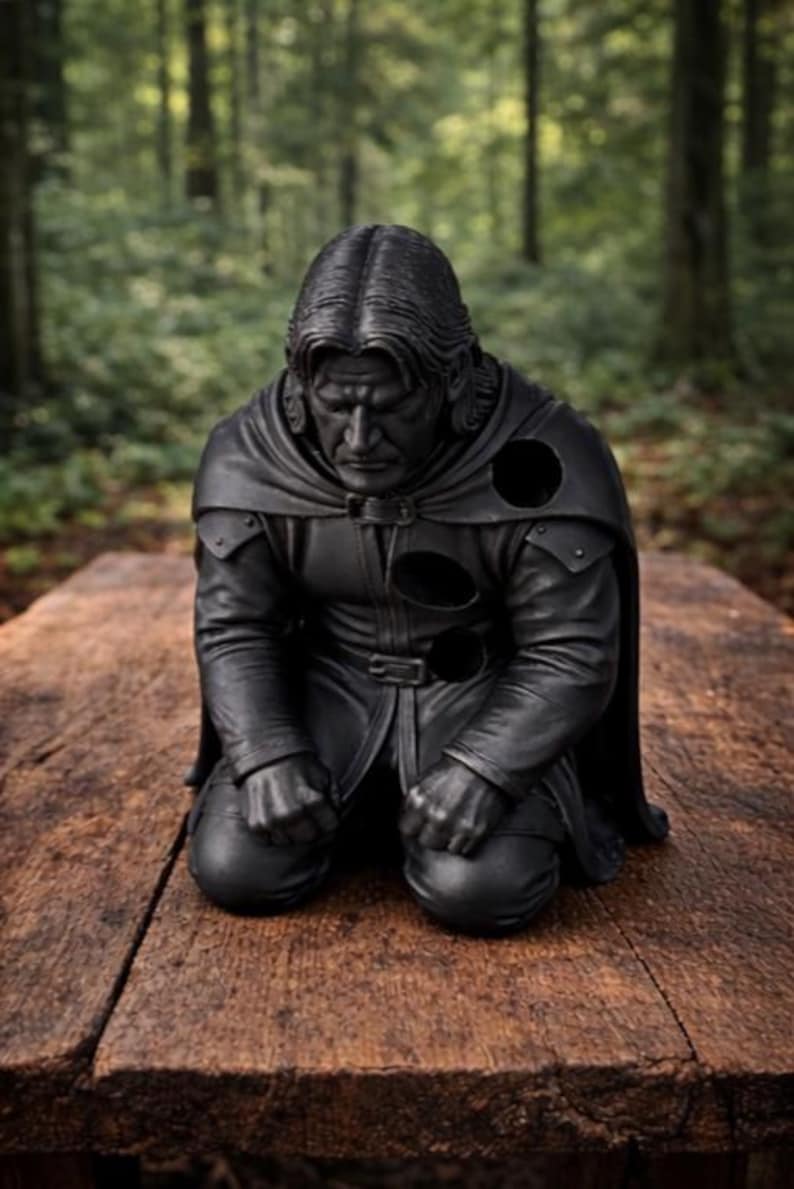 Op de afbeelding: Een zwart beeld van een knielende figuur met lang haar en een cape. Het beeld staat op een houten oppervlak, met een vage bosachtergrond. De figuur draagt een leren outfit met een riem en pantserachtige details.