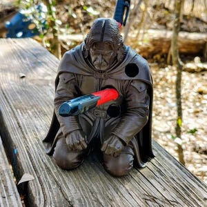 May include: A dark brown, 3D-printed figurine of a kneeling figure with long hair and a cape. The figure features circular openings designed to hold writing instruments, with a red and black pen inserted into the chest and another in the back.