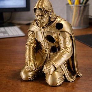 May include: A gold-colored figurine of a kneeling figure with long hair, wearing a cape and tunic. The figure has three distinct circular holes on the chest and shoulder area, with a somber facial expression and hands resting on the knees.