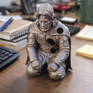 May include: A metallic bronze-colored figurine of a kneeling person with long hair, wearing a cloak and tunic. The figure features three circular holes on the chest and shoulder area, with a somber facial expression and hands resting on the knees.