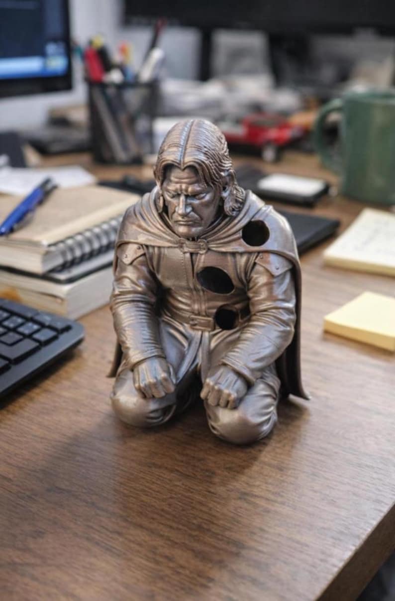 May include: A metallic bronze-colored figurine of a kneeling person with long hair, wearing a cloak and tunic. The figure features three circular holes on the chest and shoulder area, with a somber facial expression and hands resting on the knees.