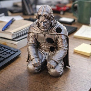 Op de afbeelding: Een bronzen beeldje van een knielende figuur met lang haar en een cape. De figuur heeft een strenge uitdrukking en draagt een gedetailleerde outfit. Het beeldje staat op een houten bureau, met kantoorbenodigdheden op de achtergrond.