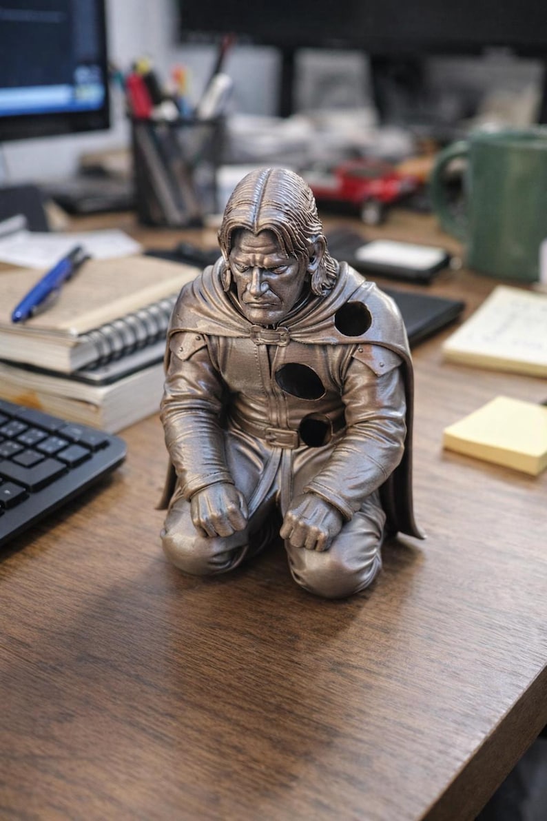 Op de afbeelding: Een bronzen beeldje van een knielende figuur met lang haar en een cape. De figuur heeft een strenge uitdrukking en draagt een gedetailleerde outfit. Het beeldje staat op een houten bureau, met kantoorbenodigdheden op de achtergrond.