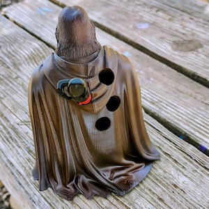 May include: A dark brown, 3D-printed figurine of a hooded figure wearing a cloak with vertical folds. The back of the cloak features three circular holes and a small, attached black and red mechanical component near the shoulder area.