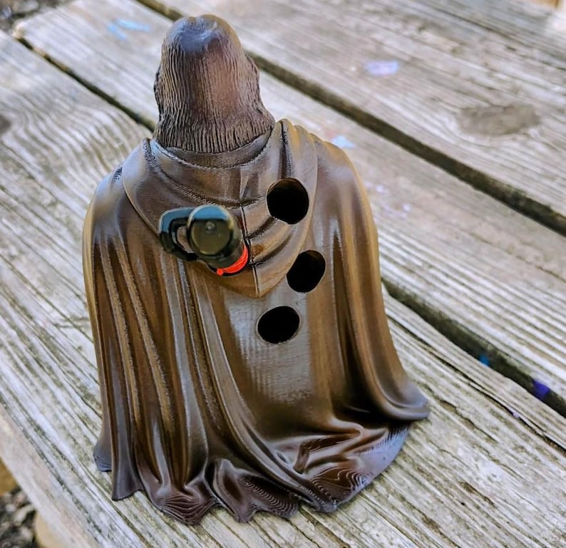 May include: A dark brown, 3D-printed figurine of a hooded figure wearing a cloak with vertical folds. The back of the cloak features three circular holes and a small, attached black and red mechanical component near the shoulder area.