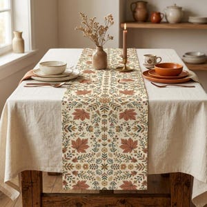 May include: A rectangular table runner featuring a repeating pattern of autumn leaves, acorns, and small flowers in shades of rust, sage green, and cream. The runner is centered on a plain beige tablecloth, accompanied by ceramic bowls and a candle.