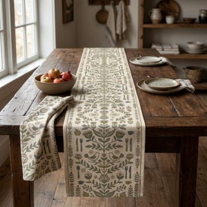 May include: A long, rectangular table runner featuring a repeating pattern of stylized birds, flowers, leaves, apples, and cutlery in muted green, brown, and cream tones. The fabric has a symmetrical design and hangs over the edge of a wooden table.