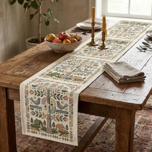 May include: A cream-colored fabric table runner featuring a repeating folk-art pattern of stylized trees, birds, and small houses in muted tones of blue, green, orange, and brown, laid across a rustic wooden dining table with a bowl of fruit.