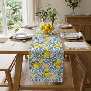 May include: A wooden dining table set for a meal, featuring a lemon-themed table runner. The runner has a white and blue pattern with yellow lemons and green leaves. The table is set with plates, glasses, and a bowl of lemons.
