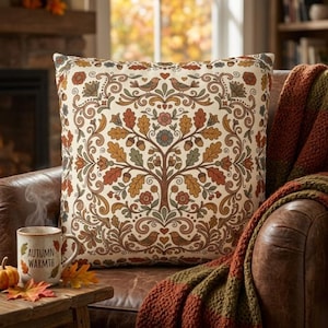 May include: A square throw pillow featuring a cream background with a symmetrical pattern of a central tree, oak leaves, acorns, flowers, and birds in shades of orange, brown, and green. The design includes small heart motifs throughout.