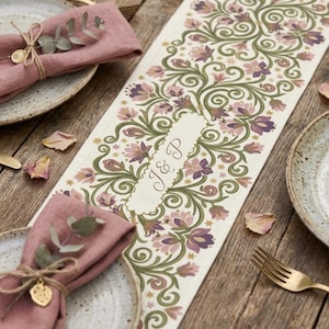 May include: A cream-coloured table runner featuring a repeating pattern of green vines and pink and purple flowers, with a central scalloped label displaying the letters J & P in cursive script, flanked by two dusty rose linen napkins with eucalyptus.