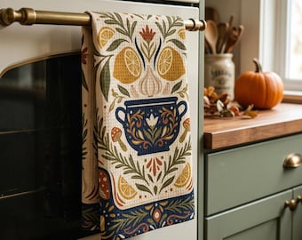 Puede incluir: Un paño de cocina de tejido de nido de abeja colgado del asa de un horno, con un estampado de una olla azul oscuro, mitades de limón amarillo, bulbos de ajo y pequeñas setas rodeadas de follaje verde sobre un fondo de color crema.