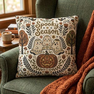 May include: A square throw pillow with a cream background featuring the text Spooky Season in brown script. The design includes three ghosts, a central jack-o'-lantern, stylized trees, mushrooms, and floral patterns in shades of brown and orange.