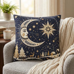Puede incluir: Un cojín cuadrado con fondo azul marino oscuro y un diseño celestial en dorado y crema. El patrón incluye una luna creciente con rostro, una estrella grande de ocho puntas, estrellas pequeñas, constelaciones, pinos, un ciervo y casas.