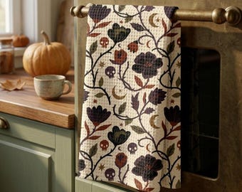 Puede incluir: Un paño de cocina de tejido gofrado con un patrón repetitivo de flores oscuras, enredaderas, pequeñas calaveras, lunas crecientes y estrellas en tonos negro, marrón y naranja apagado sobre un fondo color crema, colgado de un tirador de horno.