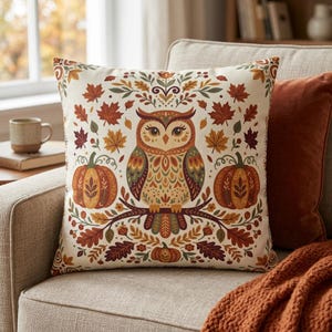 Könnte beinhalten: Ein quadratisches Dekokissen mit einer zentralen Eulenillustration auf einem Ast, flankiert von zwei orangefarbenen Kürbissen. Das Design zeigt herbstliche Motive wie Ahornblätter, Eicheln und Laub in den Farben Orange, Braun und Grün.