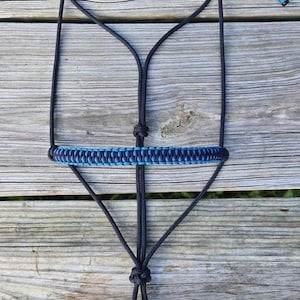 May include: A black and blue rope horse halter. The halter features a woven blue and light blue browband. The halter is made of durable rope and is designed for equine use. The halter is displayed on a wooden surface.