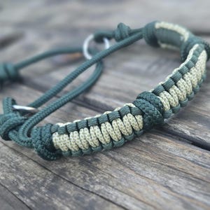 May include: A dark green and tan paracord dog collar with metal rings. The collar features a woven design in alternating colors. The background is a weathered wooden surface.