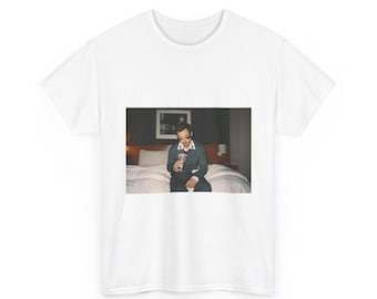 May include: White t-shirt featuring a photograph of a person sitting on a bed, holding a drink. The person is wearing sunglasses and a dark suit. The background includes a bed, a picture frame, and a lamp.