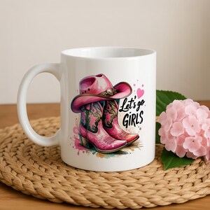 May include: A white ceramic mug featuring a graphic illustration of a pink cowboy hat and matching pink cowboy boots with decorative patterns. The text "Let's go GIRLS" is printed in black script next to a small pink heart, with colourful paint splatters.