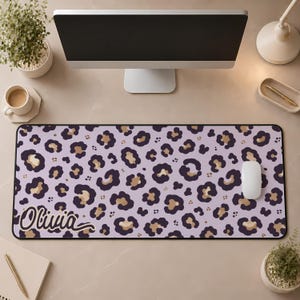 May include: A rectangular desk mat featuring a light purple background with a repeating dark purple and tan leopard print pattern. The name Olivia is printed in a dark purple cursive script in the bottom left corner of the mat.
