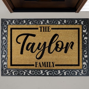 Puede incluir: Felpudo rectangular con borde negro y adornos de volutas. El centro presenta el texto "THE Taylor FAMILY" en escritura negra y letras de imprenta sobre un fondo beige. Un artículo de decoración del hogar acogedor.