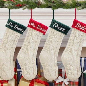 May include: Four cream-colored Christmas stockings with cable knit design hang from a mantel. The cuffs are green or red, embroidered with "Mr. Brown", "Mrs. Brown", "Groom", and "Bride" in white script. Presents and greenery decorate the scene.