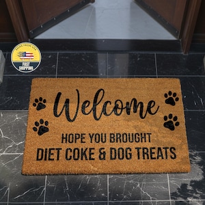 Puede incluir: Una alfombra de puerta rectangular hecha de fibra de coco marrón natural. La alfombra presenta la palabra "Welcome" en una fuente cursiva, junto con la frase "HOPE YOU BROUGHT DIET COKE & DOG TREATS". Huellas de patas negras en la alfombra.
