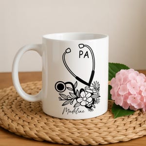 May include: White ceramic mug featuring a black stethoscope design with the letters "PA" and floral accents. The name "Madeline" is written in script below the design. The mug has a white handle.