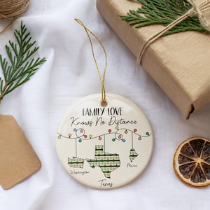 May include: A round, cream-colored ceramic ornament with the text "FAMILY LOVE Knows No Distance." The ornament features state outlines of Texas, Washington, and Maine, with a string of colorful lights. A gold string is attached for hanging.