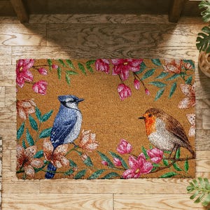 May include: A rectangular doormat featuring a blue jay and a robin perched on flowering branches. The birds are surrounded by pink and peach blossoms and green leaves against a brown background. The mat is made of a natural fiber material.