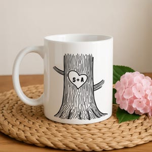 Peut inclure: Un mug en céramique blanche orné d'une illustration au trait noir représentant un tronc d'arbre avec un cœur gravé au centre. À l'intérieur du cœur, les lettres S et A sont séparées par un signe plus. Le mug a une finition lisse et une anse.