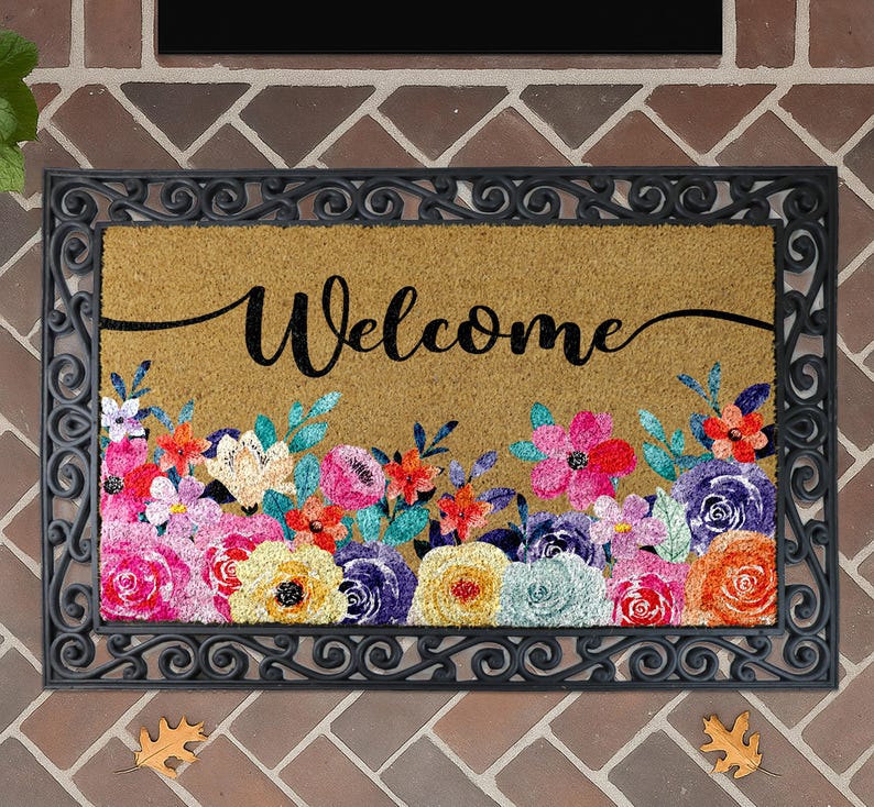 A decorative welcome mat with a floral design and the word "Welcome" written on it, placed on a brick wall.