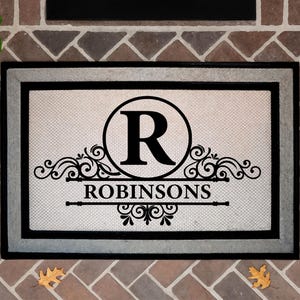 May include: Rectangular doormat with a black and gray border. The center features a large black letter "R" inside a circle, with the name "ROBINSONS" below, and decorative scrollwork. The mat is on a brick surface.