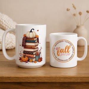 Könnte beinhalten: Zwei weiße Keramikbecher mit Herbstmotiven. Ein Becher zeigt einen Stapel Bücher mit einem Kürbis und Blättern. Der andere Becher hat ein kreisförmiges Design mit dem Wort "Fall" und Wörtern wie "Blätter" und "Apfelwein".