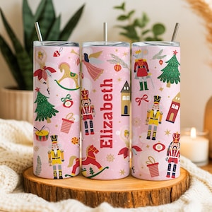 May include: Three pink insulated tumblers with a Christmas-themed design. Each tumbler features illustrations of nutcrackers, Christmas trees, and other festive elements. The name "Elizabeth" is printed vertically on one tumbler. Each tumbler has a silver straw.
