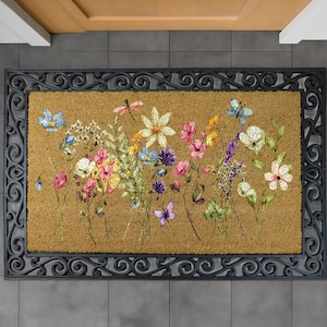 May include: Rectangular doormat with a floral design. The mat features a brown coir center with a colorful array of painted flowers and butterflies. A black, ornate border frames the design. The mat is placed on gray tile flooring.