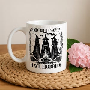 May include: A white ceramic mug featuring a black graphic illustration of three witches standing around a campfire with the text "GOD FORBID WOMEN HAVE HOBBIES" printed in a bold, serif font above and below the central witch design.