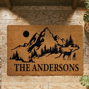 A wooden doormat with a mountain landscape and deer silhouette, along with the text "THE ANDERSONS" printed on it.