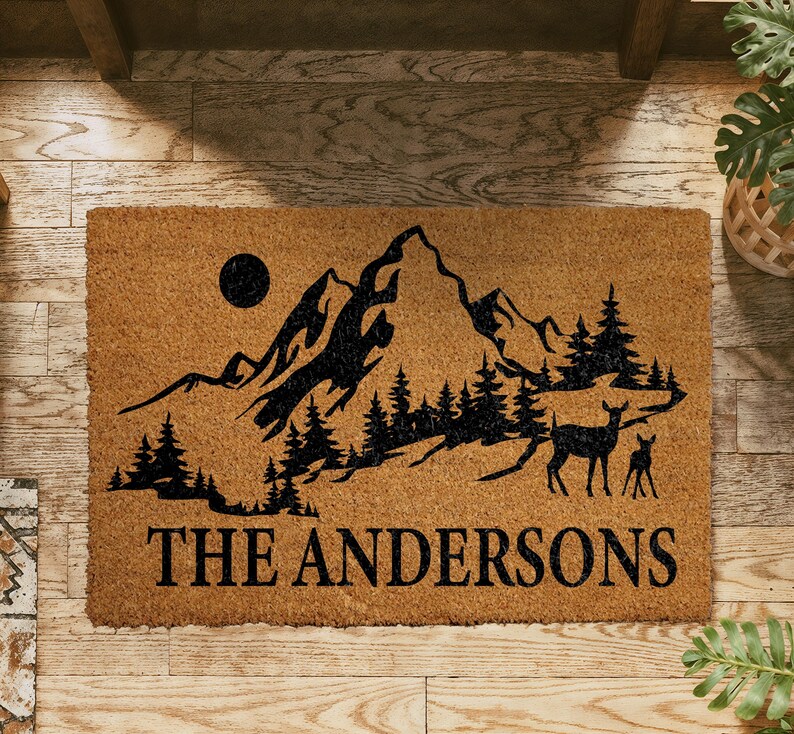 A wooden doormat with a mountain landscape and deer silhouette, along with the text "THE ANDERSONS" printed on it.