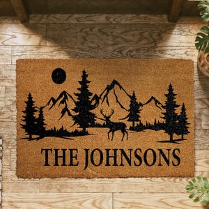 A wooden doormat with a mountain landscape and a deer silhouette, along with the text "THE JOHNSONS" printed on it.