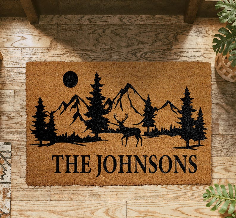 A wooden doormat with a mountain landscape and a deer silhouette, along with the text "THE JOHNSONS" printed on it.