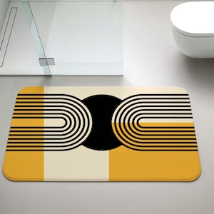 May include: A rectangular bath mat with a geometric design in a modern bathroom setting. The mat features a black circle and curved lines in black and white, set against a yellow and cream background. The mat is approximately 30 inches long.