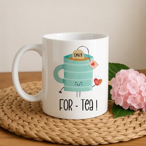 May include: A white ceramic mug featuring a cartoon illustration of a light blue striped mug with a smiling face, holding a small heart with the number 40. A tea bag tag labeled Emily hangs from the top. The text FOR - Tea! is printed below.