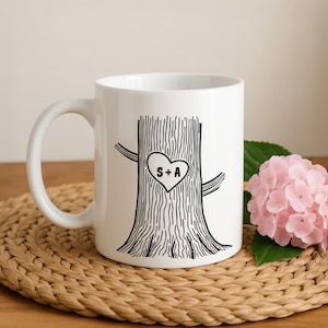 May include: White ceramic mug featuring a black tree trunk design with a heart-shaped carving. The heart contains the initials "S+A". The mug has a curved handle and is placed on a woven mat.