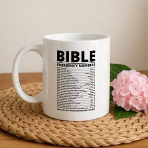May include: A white ceramic mug featuring black printed text titled BIBLE EMERGENCY NUMBERS. The text lists various life situations followed by corresponding Bible verses, ending with the phrase AVAILABLE 24/7 PHONE SERVICE NOT REQUIRED.