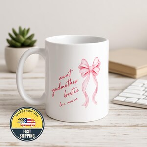 May include: A white ceramic mug featuring a pink ribbon illustration and red text that reads "aunt godmother bestie love, maria". The mug has a curved handle and a smooth finish, displaying the printed design clearly on the front surface.